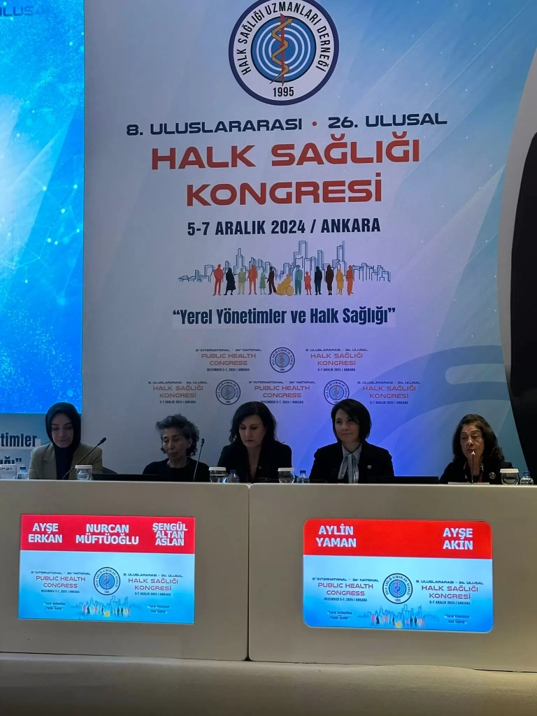 Bir kongrede çekilmiş bir panel oturumu görüntüsü. Fotoğrafın arka planında "8. Uluslararası - 26. Ulusal Halk Sağlığı Kongresi, 5-7 Aralık 2024 / Ankara" yazılı bir pano yer alıyor. Ayrıca "Yerel Yönetimler ve Halk Sağlığı" teması dikkat çekiyor. Pano üzerinde "Halk Sağlığı Uzmanları Derneği 1995" logosu bulunuyor ve görselde şehir silüetleri ve farklı insan figürleri resmedilmiş. Ön planda, oturum konuşmacıları bir panel masasında oturuyor. Masanın üzerinde konuşmacıların isimlerini gösteren dijital ekranlar yer alıyor. Soldan sağa şu isimler yazılı: Ayşe Erkan, Nurcan Müftüoğlu, Şengül Altan Arslan, Aylin Yaman ve Ayşe Akın. Konuşmacılar profesyonel bir görünüme sahip ve kürsüde mikrofonlar ile su şişeleri bulunuyor. Arka planda, panelin düzenlendiği kongre ortamını yansıtan görseller yer alıyor.